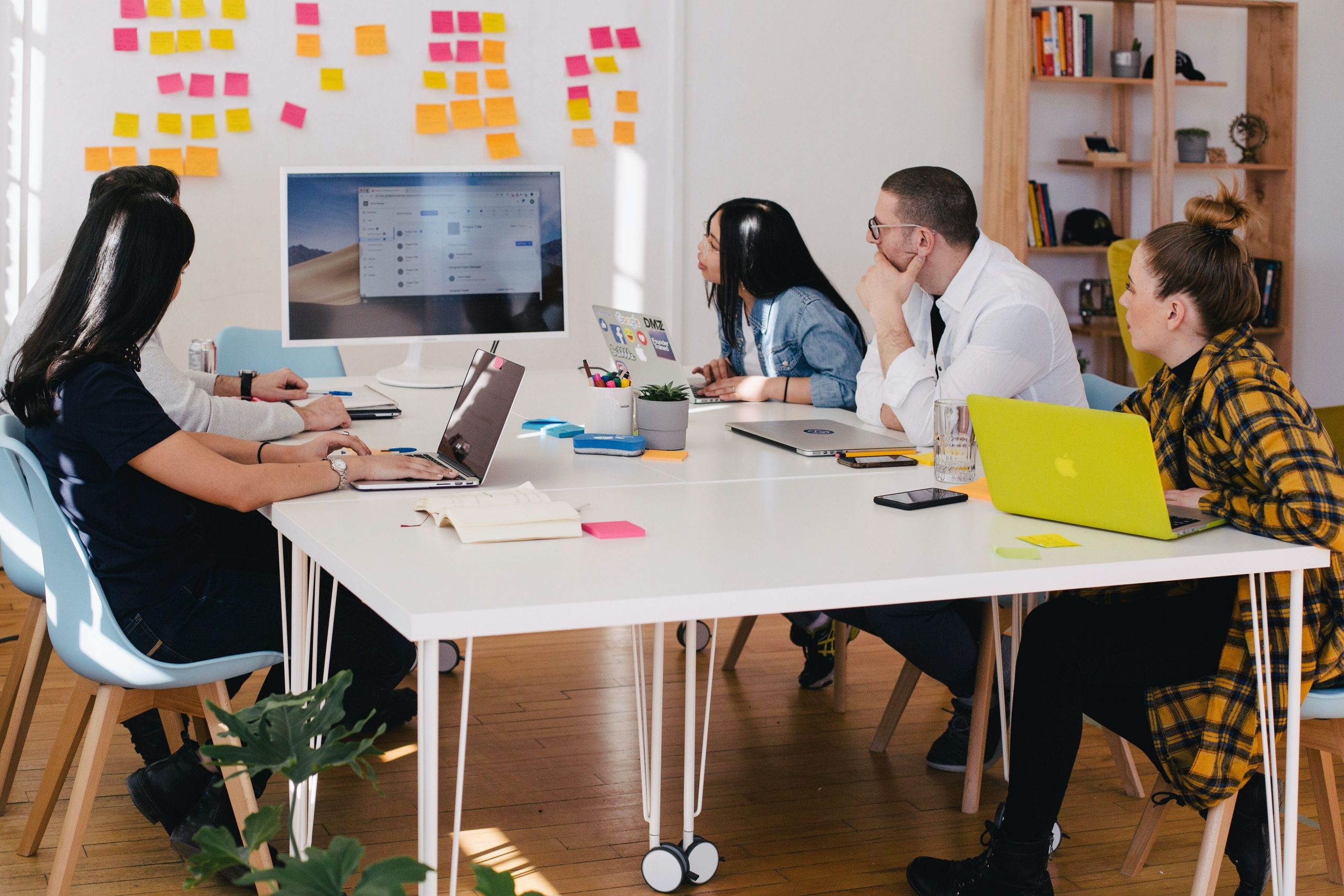 5 people sitting around a white table in a meeting together, they are looking at a screen.