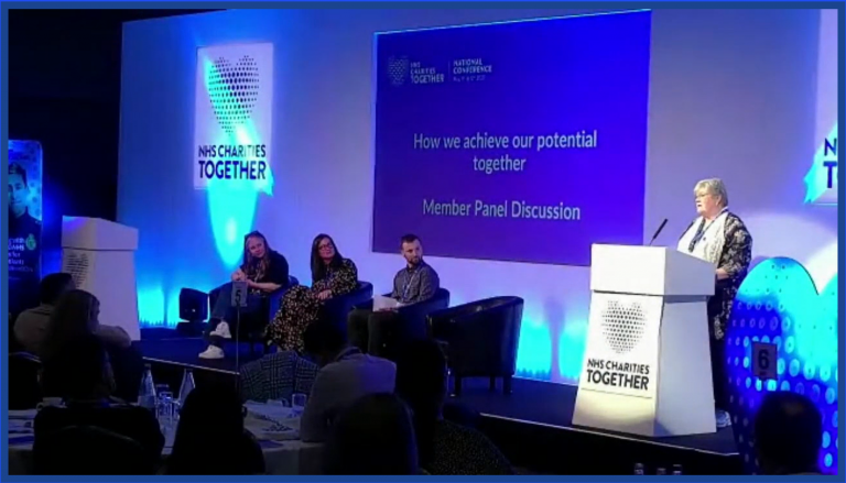 an image showcasing a panel discussion from the in-person portion of the hybrid event. There are 4 people in this image on a stage, with one stood at a podium